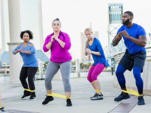 A group of diverse individuals performing leg strengthening exercises using resistance bands outdoors, demonstrating power band workouts for mobility and flexibility.