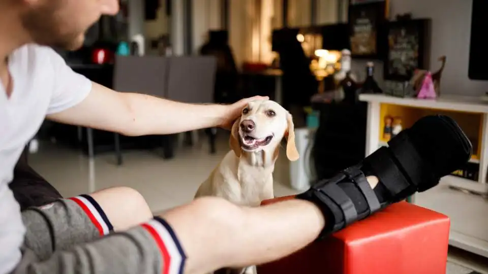 Young man with an orthopaedic boot on his right leg is petting his dog.