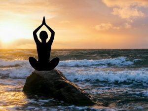Silhouette of a person practicing yoga breathing techniques for relaxation on a rock by the ocean during sunset, showcasing calm and mindfulness