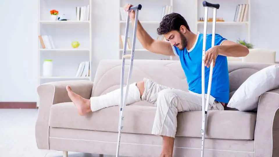Young male trying to sit on the sofa. He has a broken leg and uses crutches for first time.