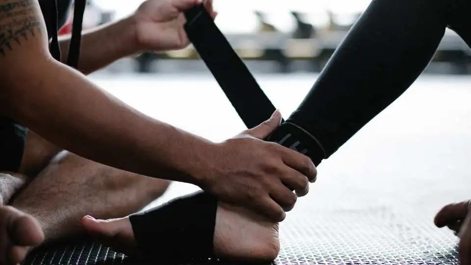 A man helping fellow gym friend with placing an elastic ankle support the right way.
