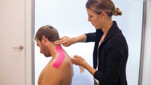 Physio applying pink kinesio tape on a patient's neck to support his traps, neck and shoulder movement.