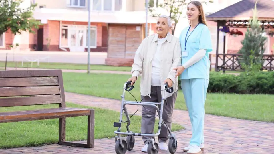 Carer helping an elderly person to walk with a four-wheeled rollator in the park.