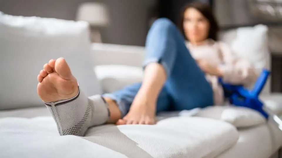 Young woman resting her foot on the sofa while using an elastic ankle support to control the swelling and inflammation.