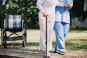 An elderly person using a walking cane assisted by a caregiver outdoors, with a wheelchair in the background, highlighting the difference between canes and walking sticks
