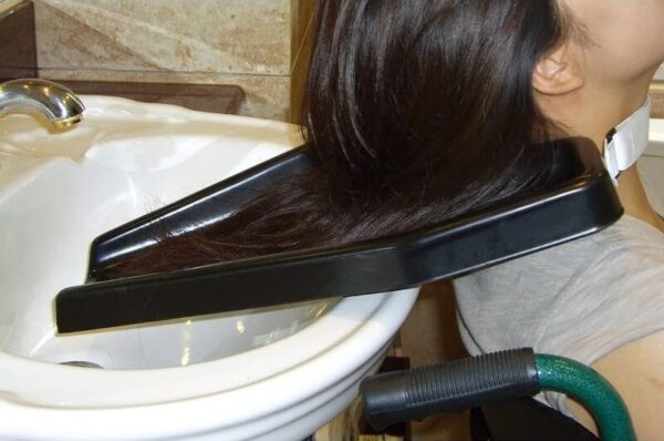 Woman using a black hair wash tray positioned between her neck and a sink, showing how the tray directs water safely into the basin