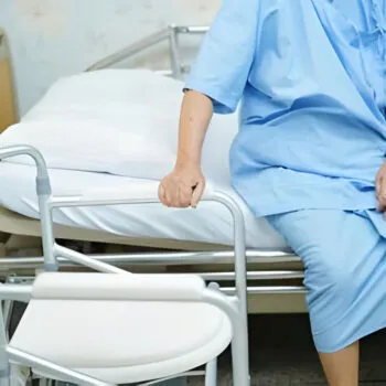 An elderly person sitting on a hospital bed beside a folding commode chair, highlighting the features of medical and heavy-duty commode chairs for home and hospital use.