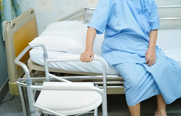 An elderly person sitting on a hospital bed beside a folding commode chair, highlighting the features of medical and heavy-duty commode chairs for home and hospital use.